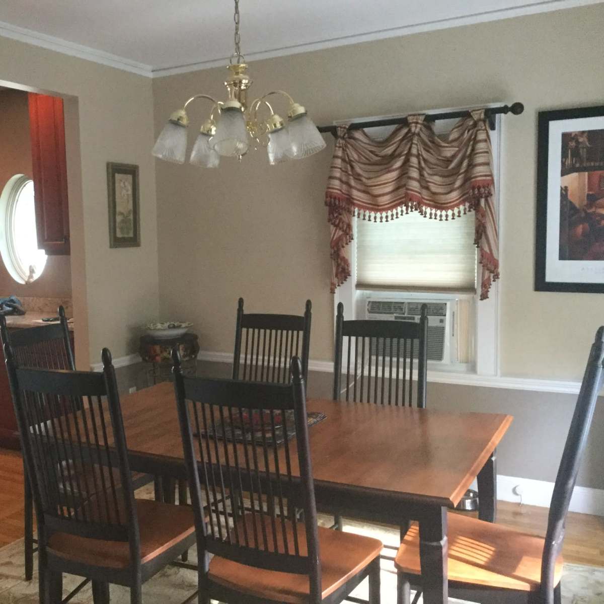 A dining room features a wooden table surrounded by black chairs, with a view of a kitchen in the background.