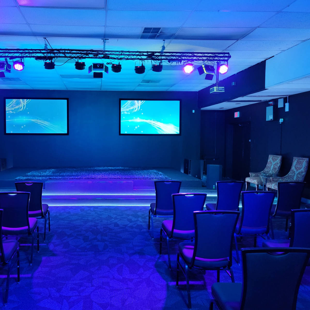A stage area equipped with two screens and purple lighting, surrounded by rows of chairs.