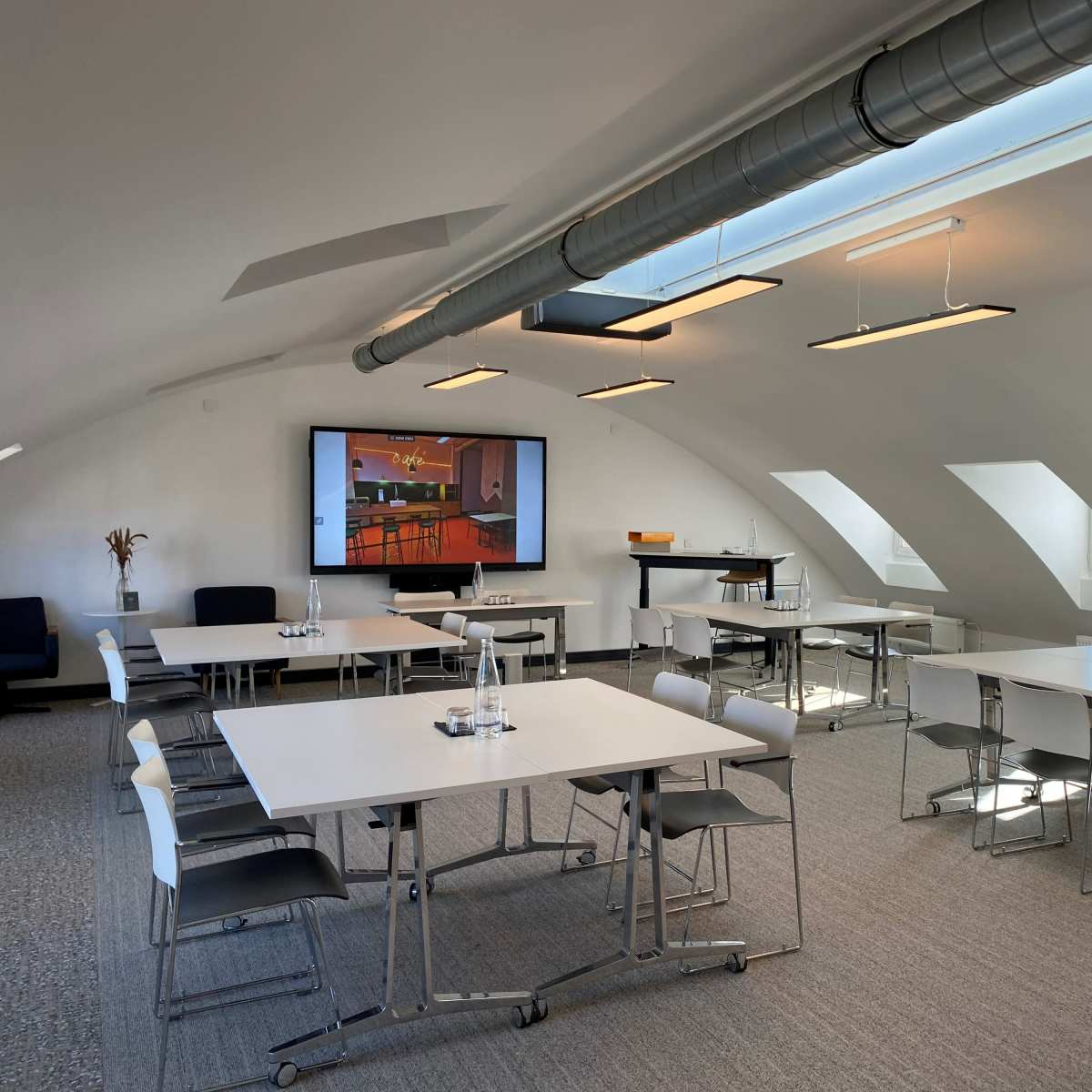 A modern meeting room features multiple tables and chairs arranged around a central area with a large screen displaying a presentation.
