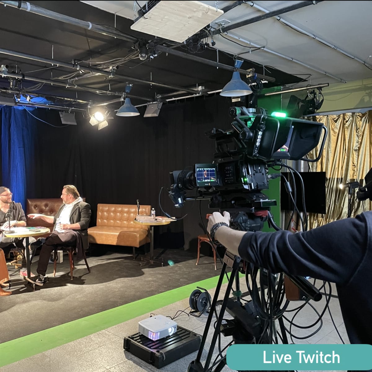 A live Twitch event is taking place on stage with a camera focused on a group of people seated and engaged in conversation.