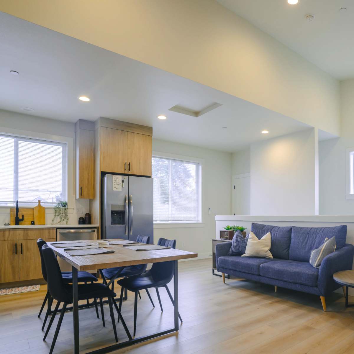 A modern, open-concept living area featuring a blue sofa, a dining table with chairs, and a kitchen with wooden cabinets.