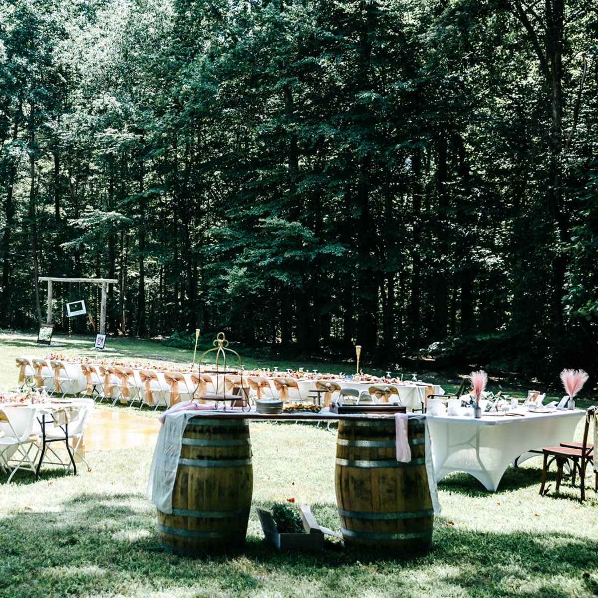 A beautifully arranged outdoor dining setup with multiple long tables, adorned with centerpieces, under a tree canopy.