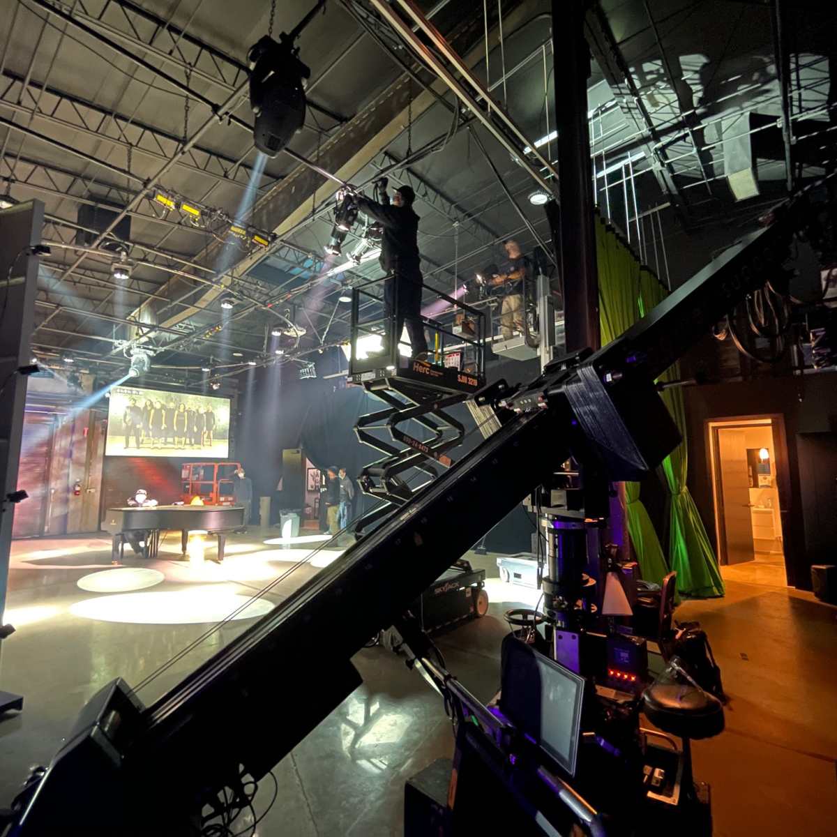 A film set is shown with crew members operating equipment on a platform above a stage illuminated by spotlighting.