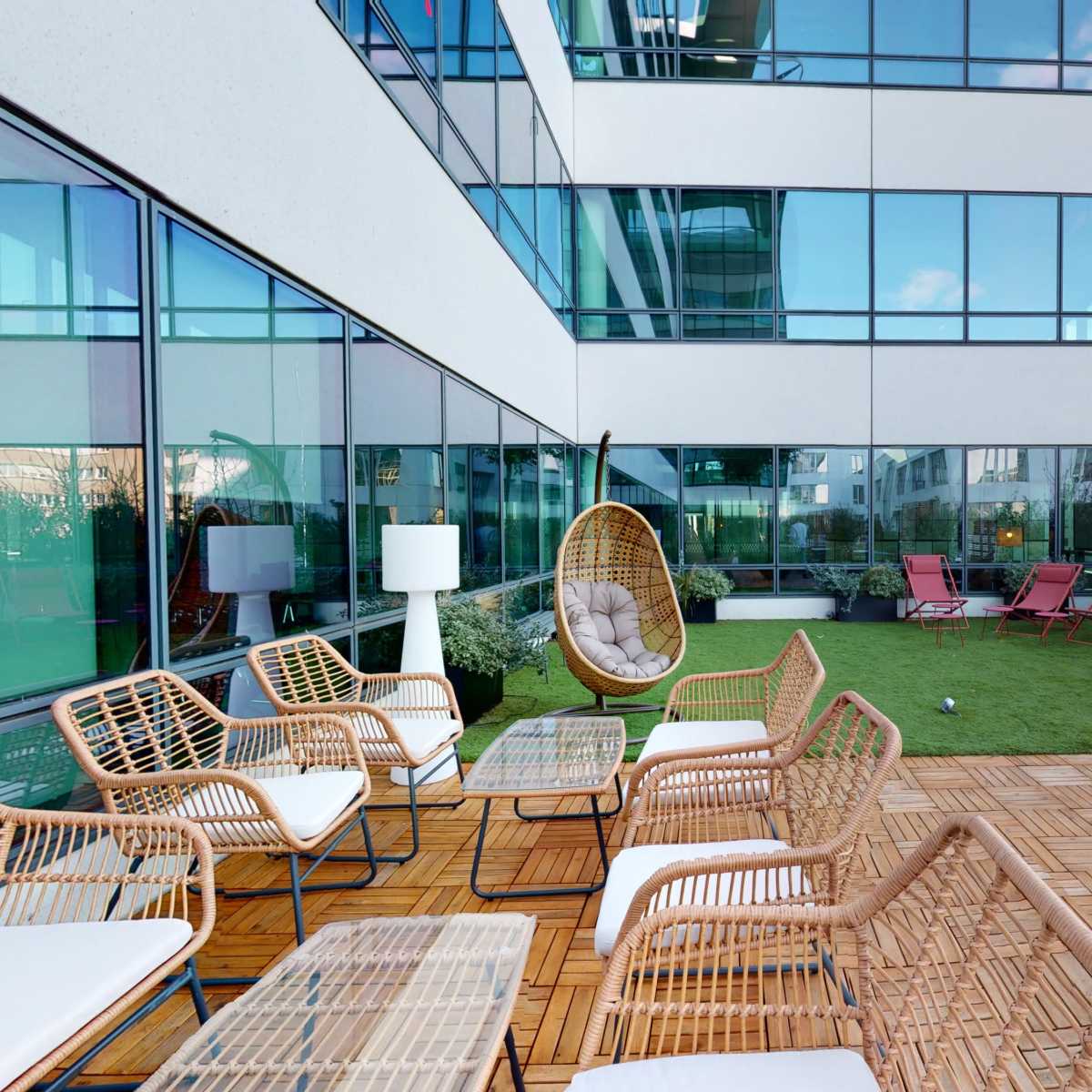 An outdoor lounge area with rattan furniture, including chairs and tables, on a wooden deck surrounded by greenery and modern glass buildings.