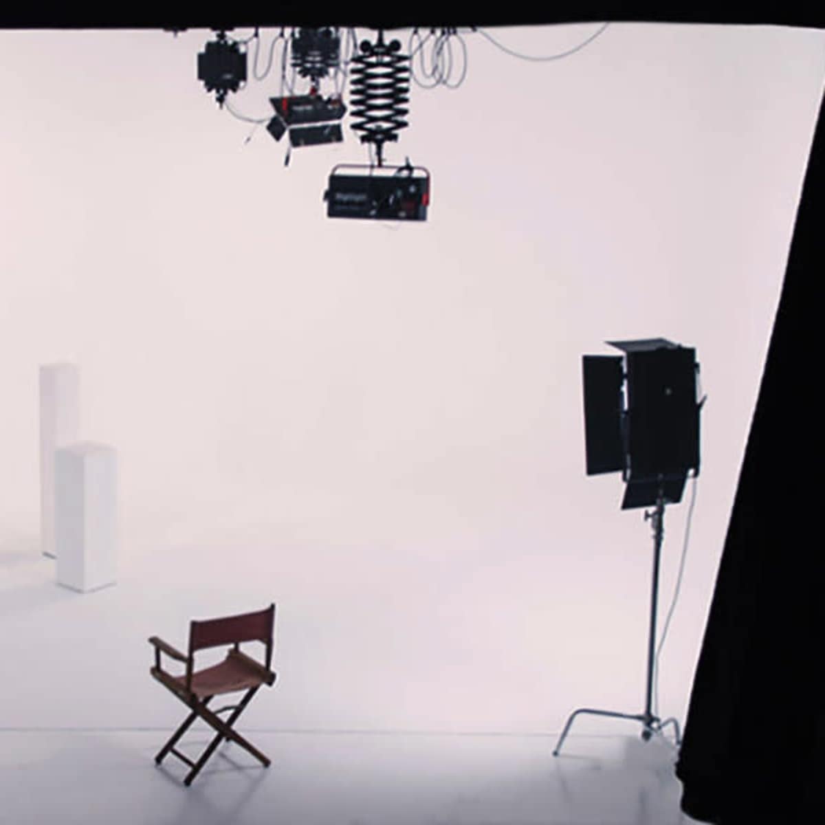 A white studio backdrop with two light stands, two light sources, and a wooden director's chair placed in front of two white pedestals.