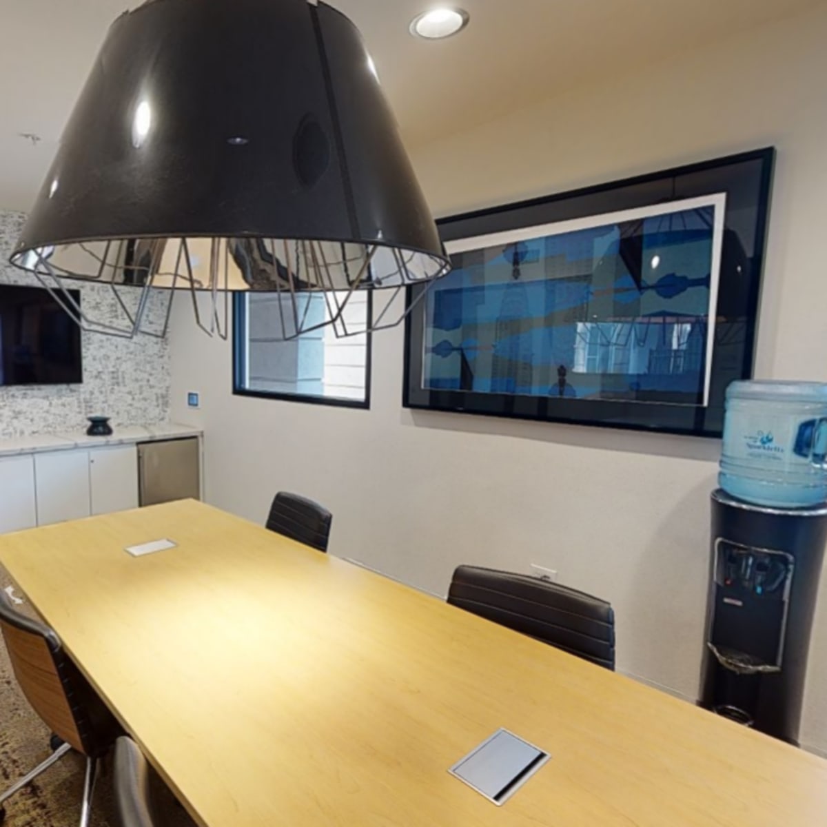 A modern conference room with a long wooden table, a large pendant light, and a water cooler against a wall with decorative artwork.