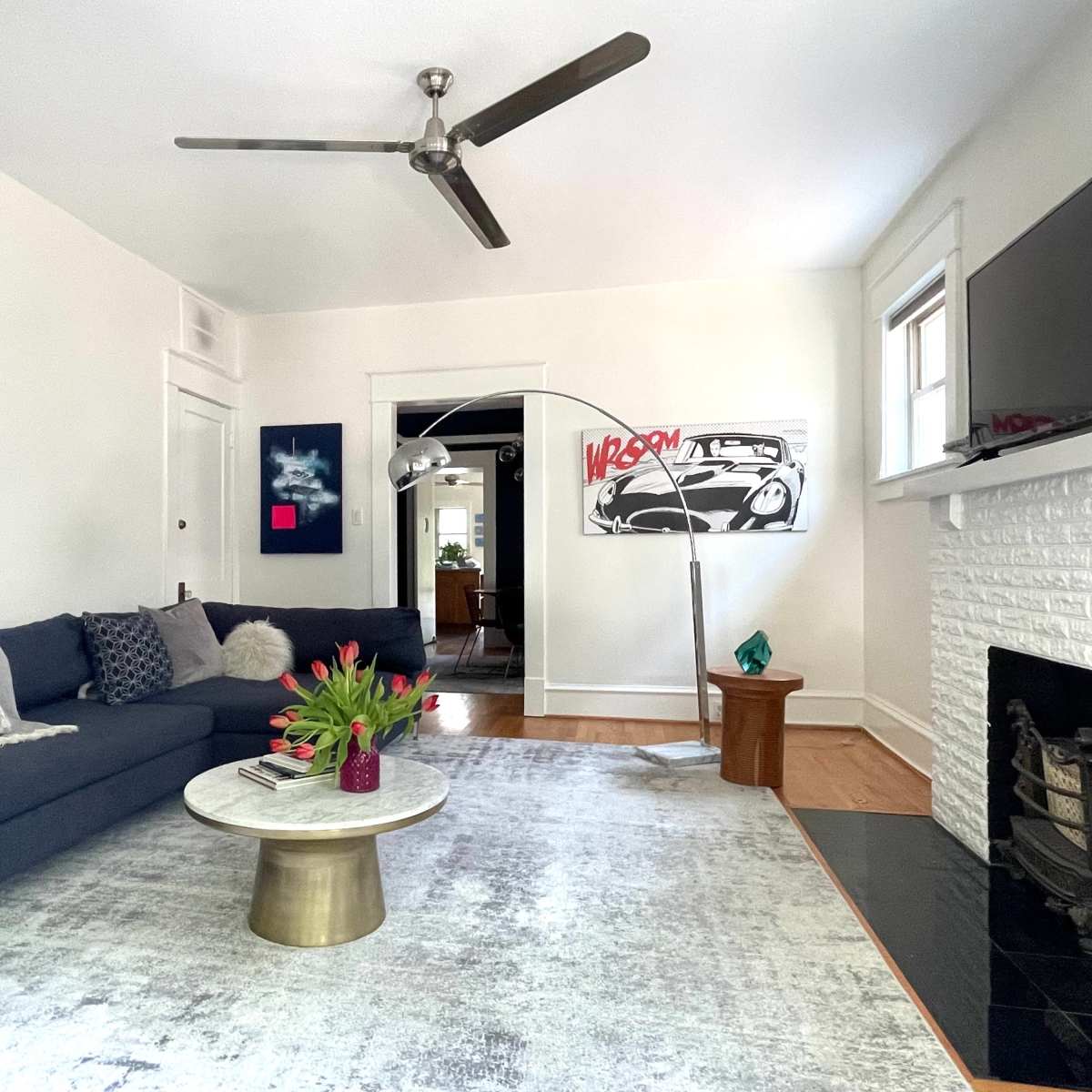 A spacious living room features a dark blue sofa, a round marble coffee table, and a white brick fireplace, with a modern ceiling fan overhead and artwork on the walls.