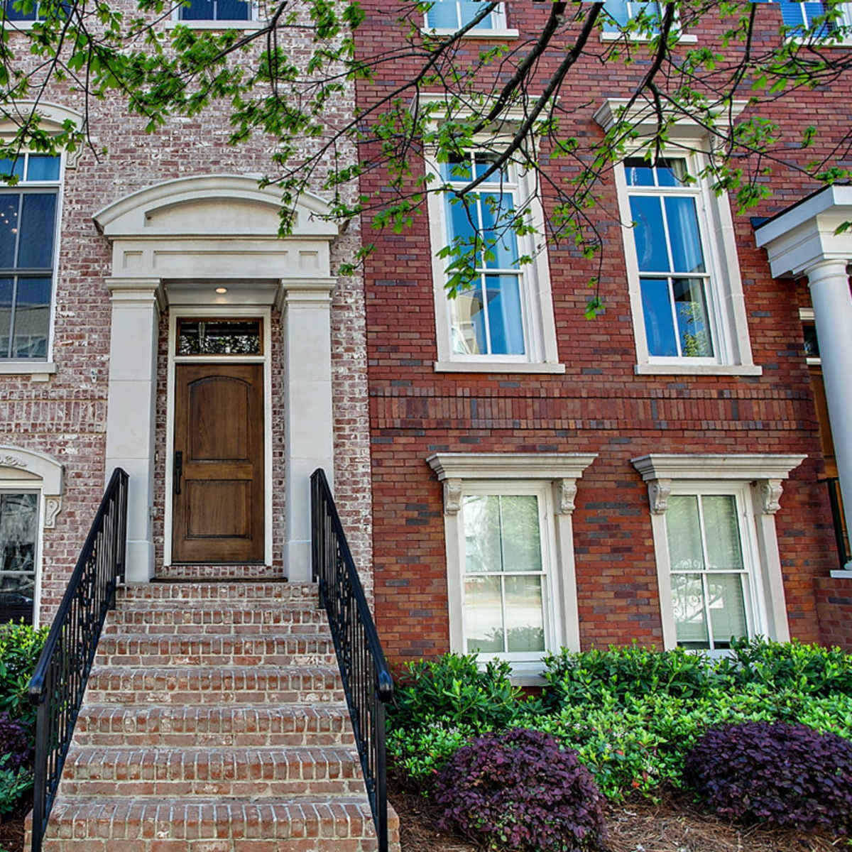 A brick townhouse with a central wooden door, flanked by tall windows and flanked by brick and greenery on either side.