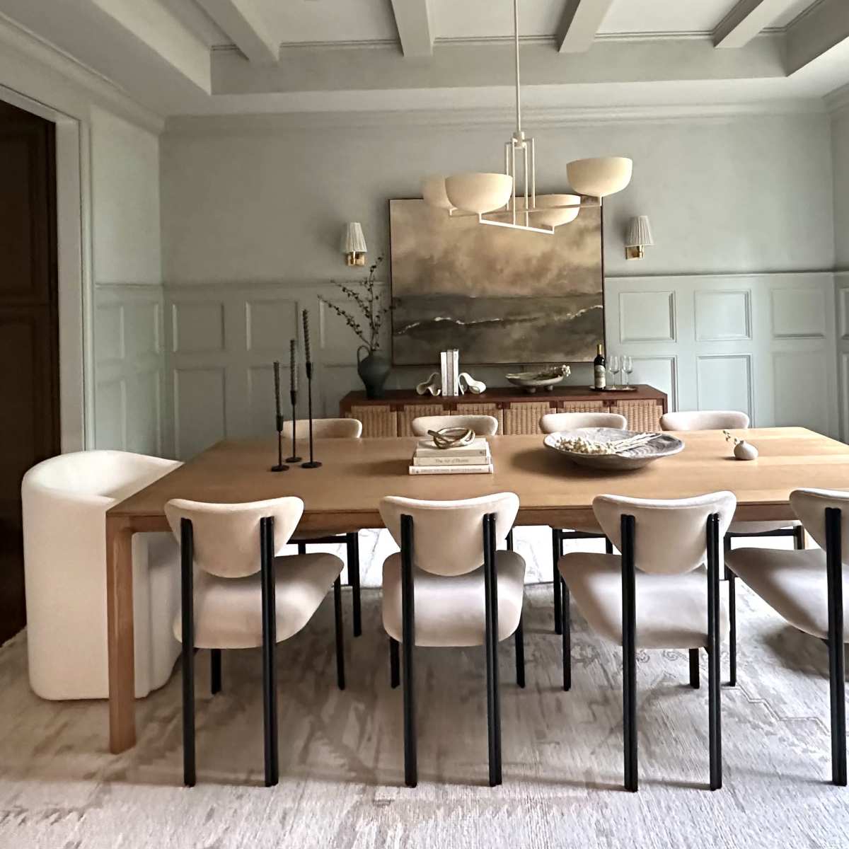 A modern dining room featuring a large wooden table surrounded by eight upholstered chairs, with a decorative centerpiece and a large artwork on the wall.