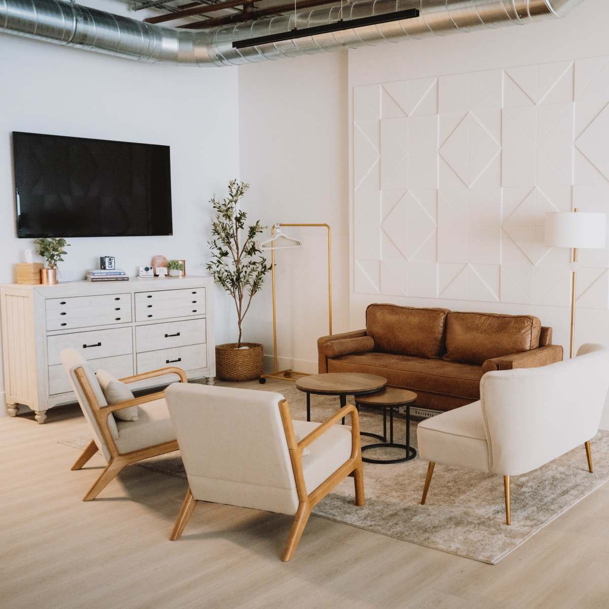 A modern living room features a brown leather sofa, two white chairs, a coffee table, a large mirror, and a TV mounted on the wall.