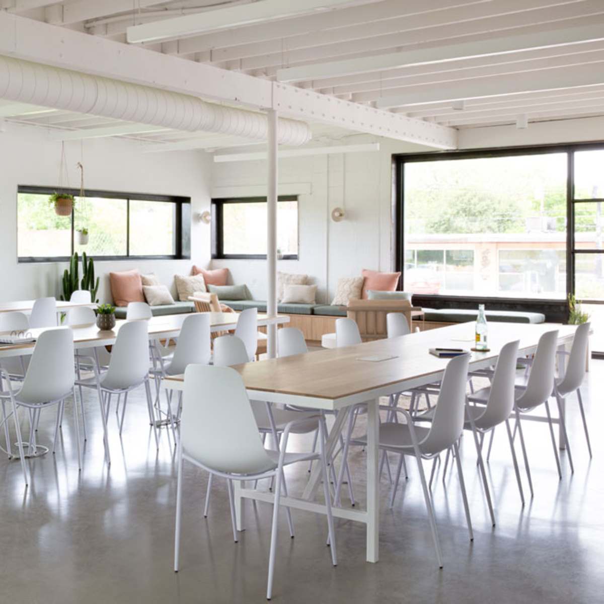 A bright, modern office space with several tables and chairs arranged for meetings, large windows allowing natural light, and a cozy seating area in the background.