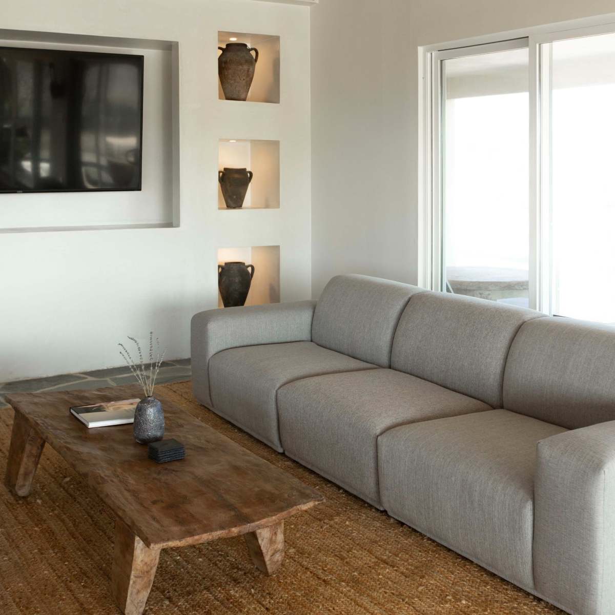 A modern living room featuring a light gray sectional sofa, a wooden coffee table, a flat-screen TV mounted on the wall, and decorative vases displayed in recessed niches.