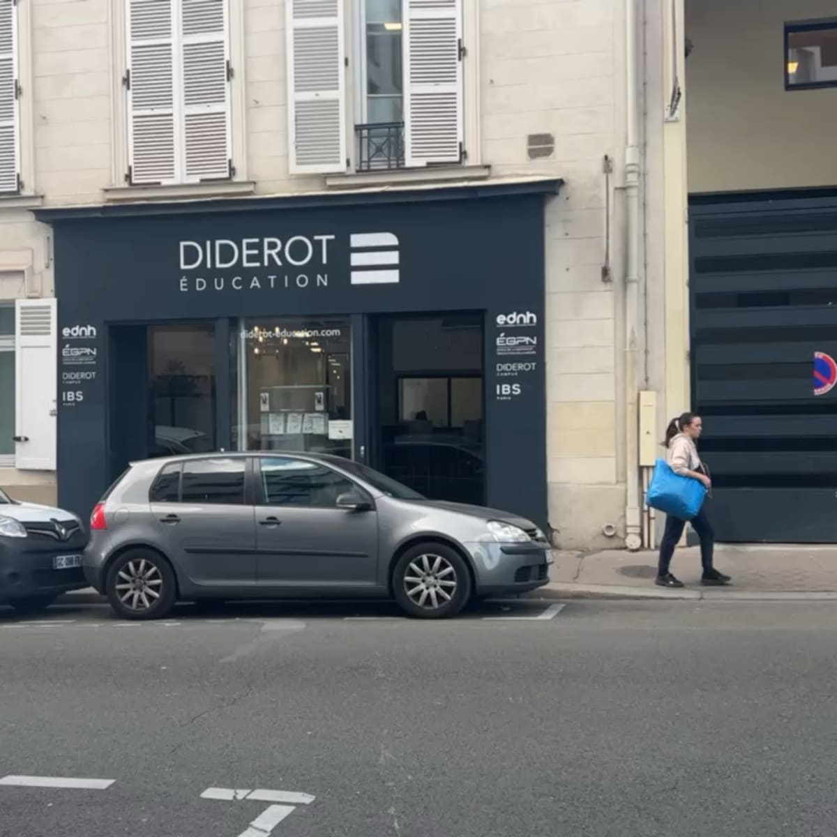 A gray car parked on the street in front of a building with a sign reading "Diderot Education" and a woman walking by carrying a blue bag.