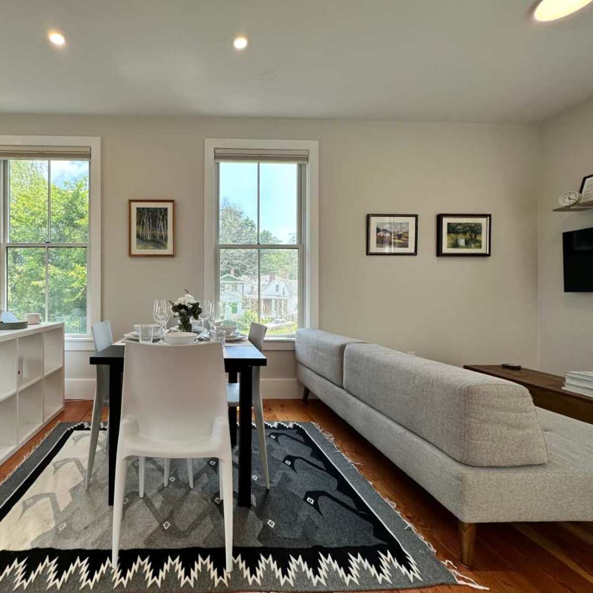 A bright, modern living space featuring a dining table set for two, a gray sofa, and a white bookshelf, with large windows allowing natural light to enter.