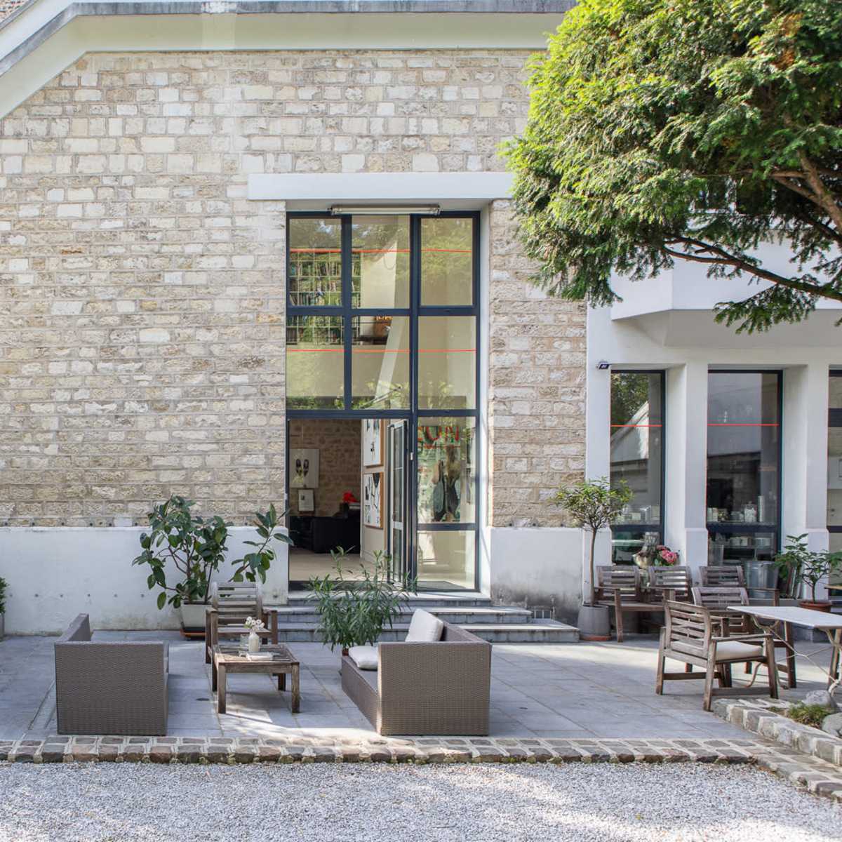 A modern stone building with large glass windows, surrounded by greenery and outdoor seating arrangements on a paved patio.