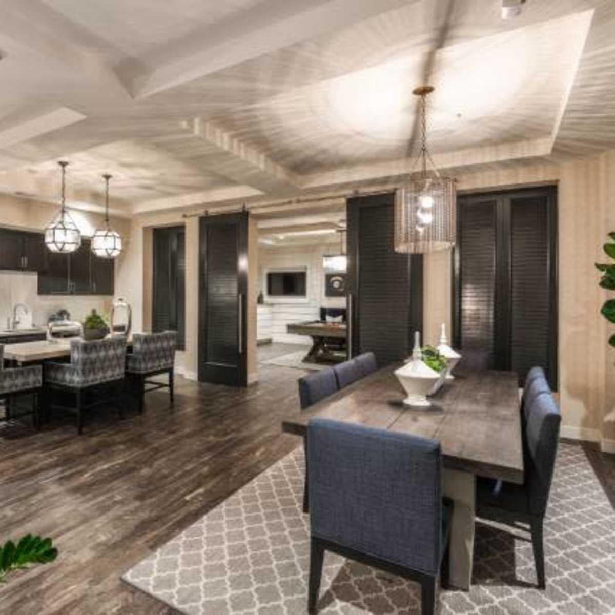 An open-concept dining and kitchen area featuring a wooden dining table, upholstered chairs, modern light fixtures, and plants.