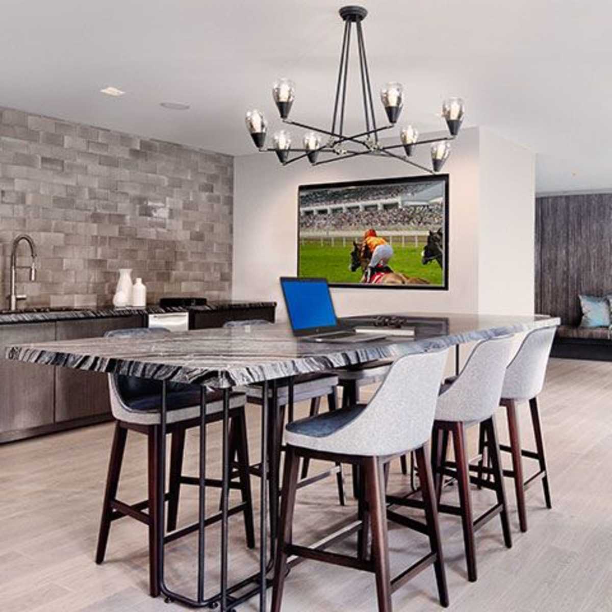 A modern kitchen or dining area with a large island, several high stools, a wall-mounted television displaying a sports event, and a cozy seating area in the background.