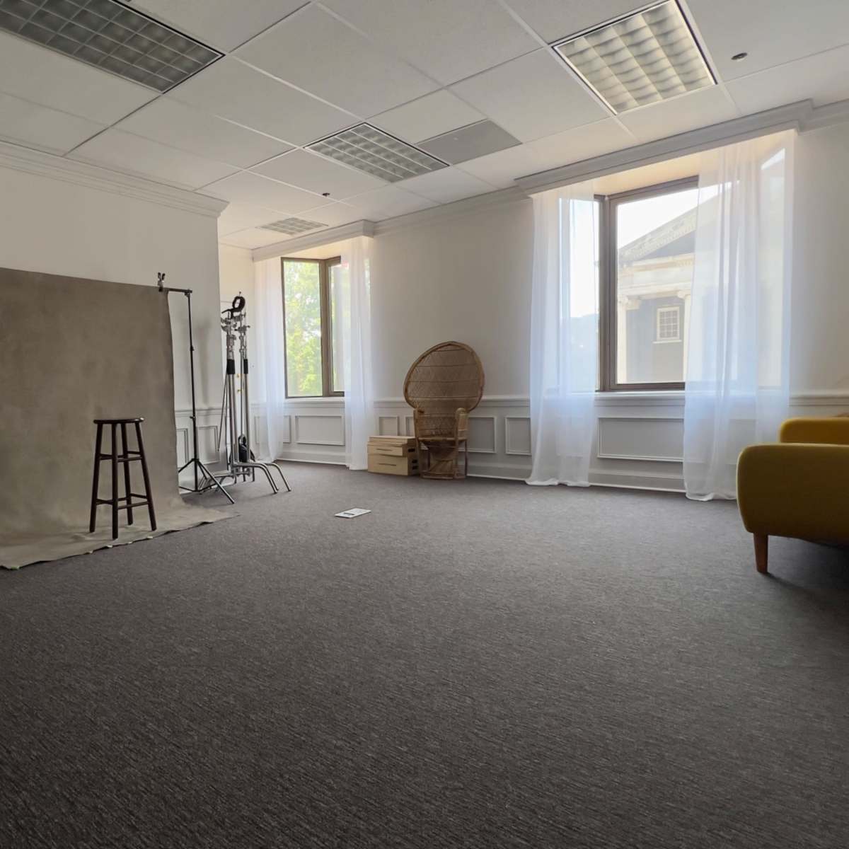 A spacious, well-lit room with a backdrop setup, a stool, and a decorative chair near large windows.