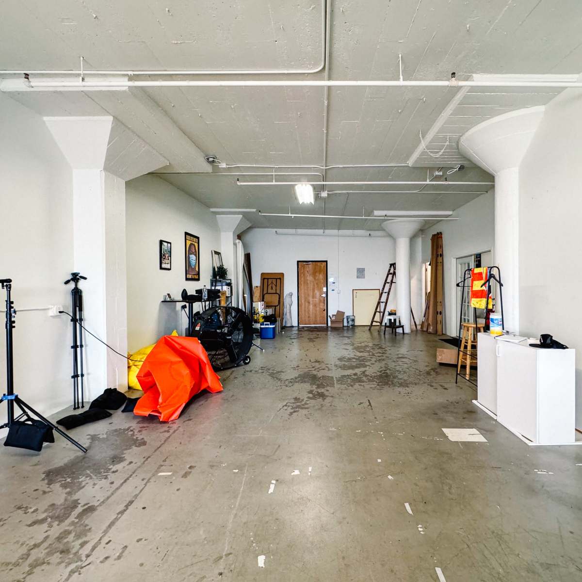 A spacious, empty room with concrete flooring, white walls, and various equipment and props arranged along one side.