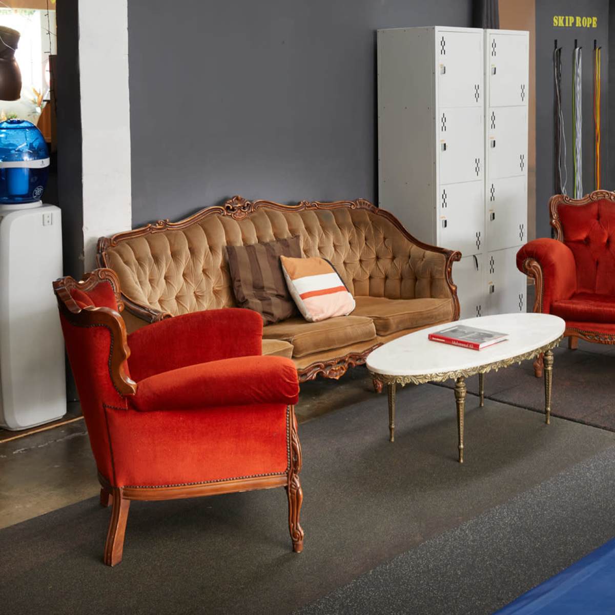 A vintage seating area with an ornate brown sofa and two red armchairs arranged around a white coffee table in a gym setting.