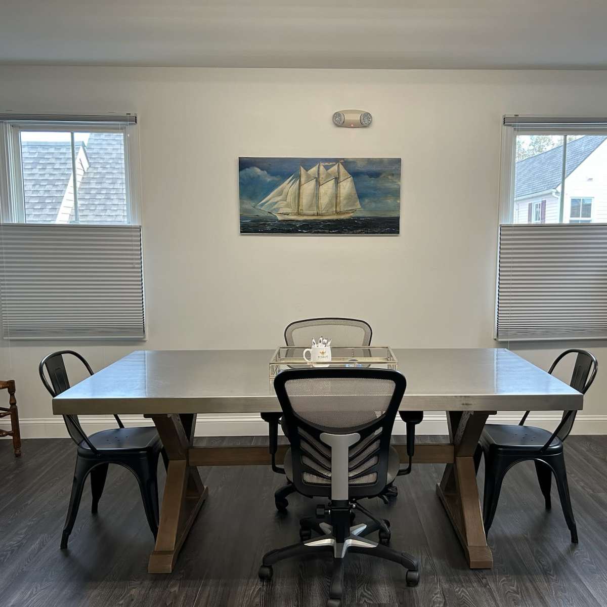 A spacious dining area features a large table with five chairs, framed by two windows with blinds and a painting of a sailing ship on the wall.