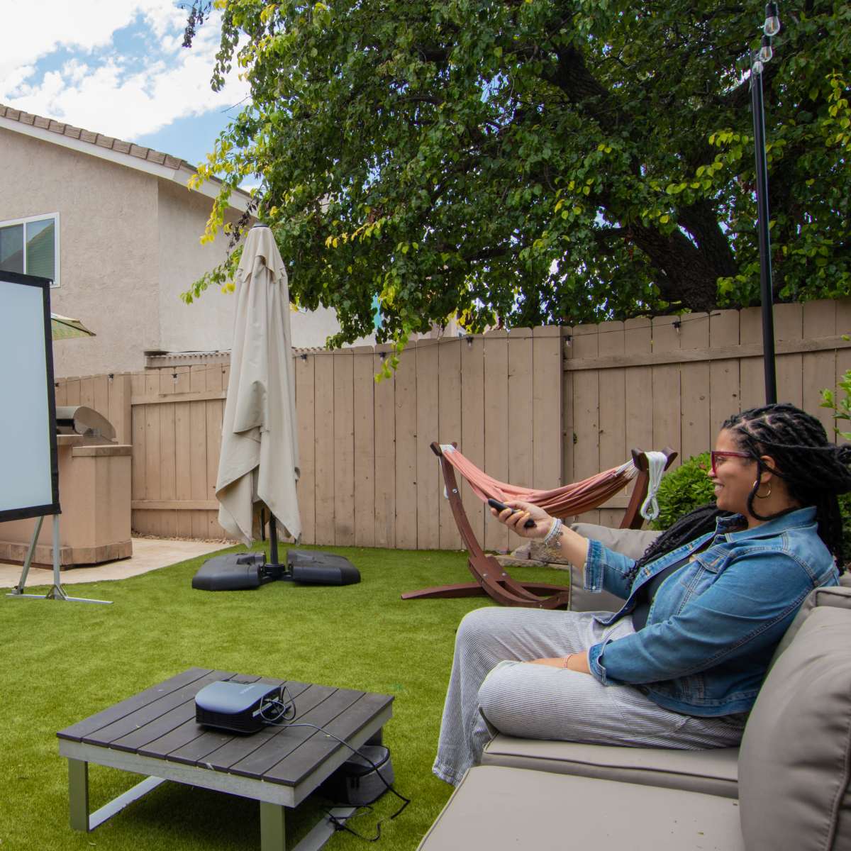 A woman sits on a sofa in a backyard, pointing a remote at a large projection screen set up on the lawn.