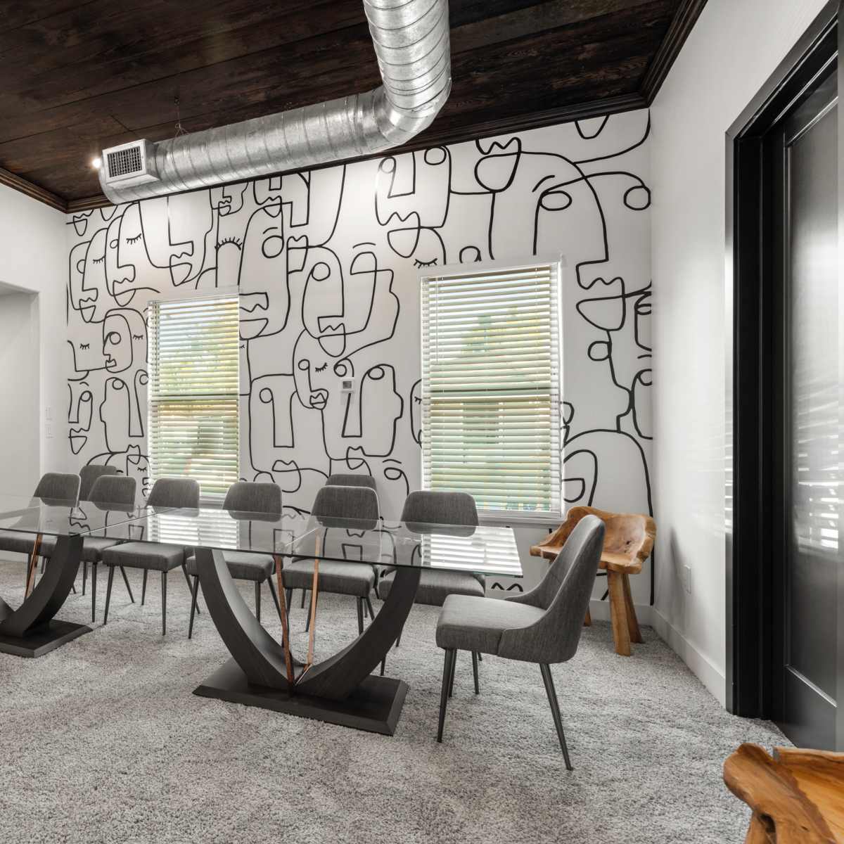 A modern conference room features a large glass table surrounded by gray chairs, with a mural of abstract line art on the wall and a wooden accent chair in the corner.