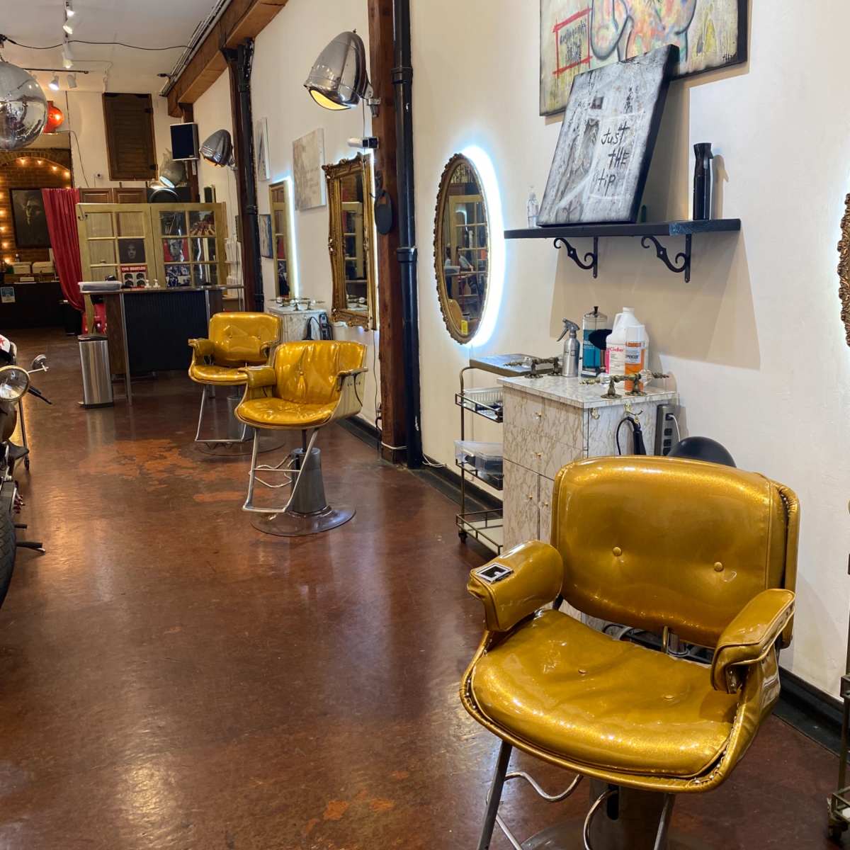 A hair salon with three vintage yellow chairs positioned in a row against a wall, with mirrors and various salon equipment in the background.