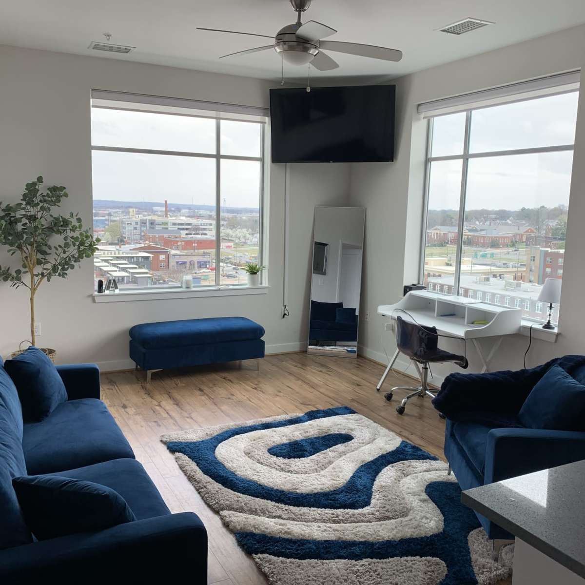 A modern living room with two blue sofas, a mirror, a desk with a chair, and large windows overlooking an urban landscape.