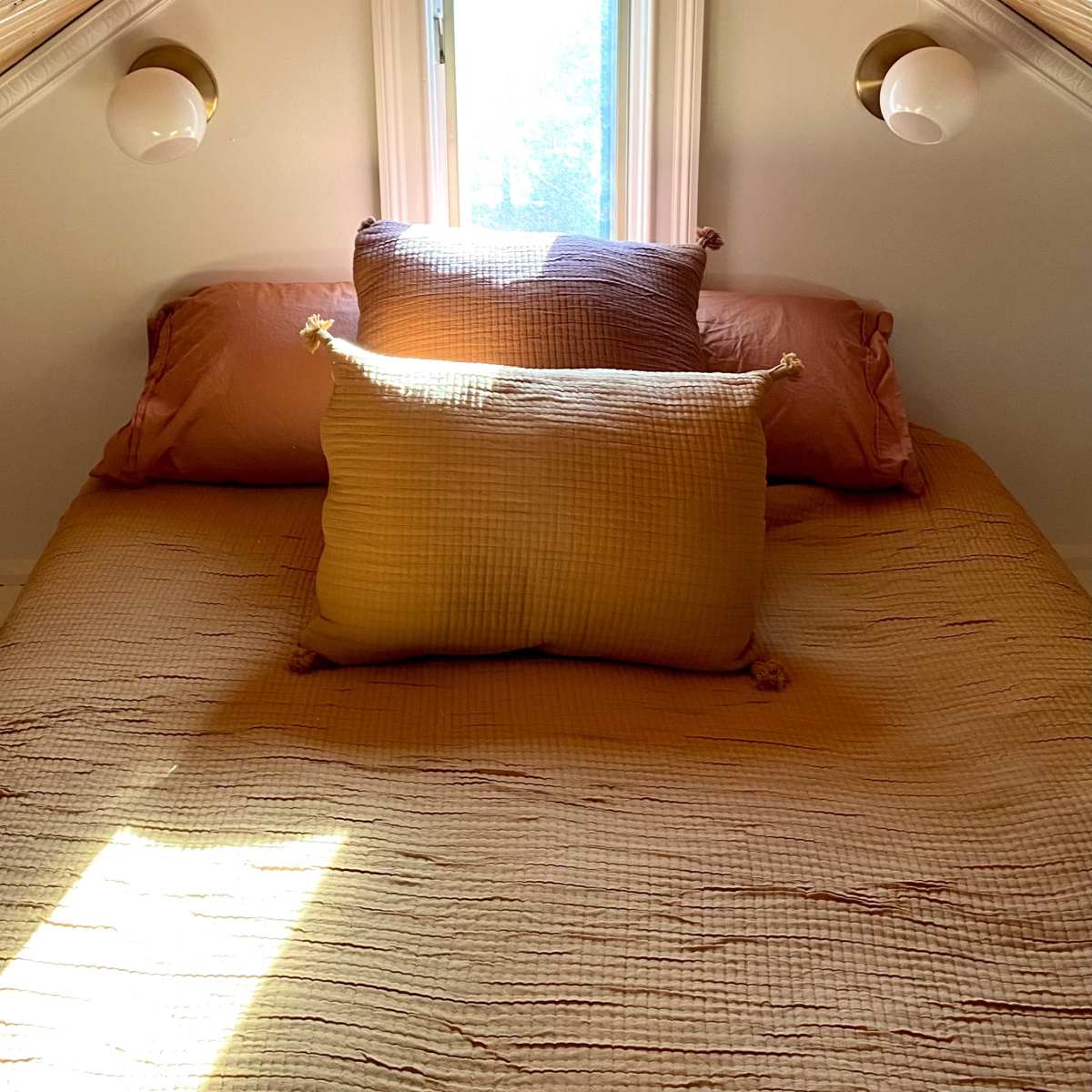A cozy small bedroom with triangular slanted walls, a window letting in light, and several pillows arranged on a neatly made bed.