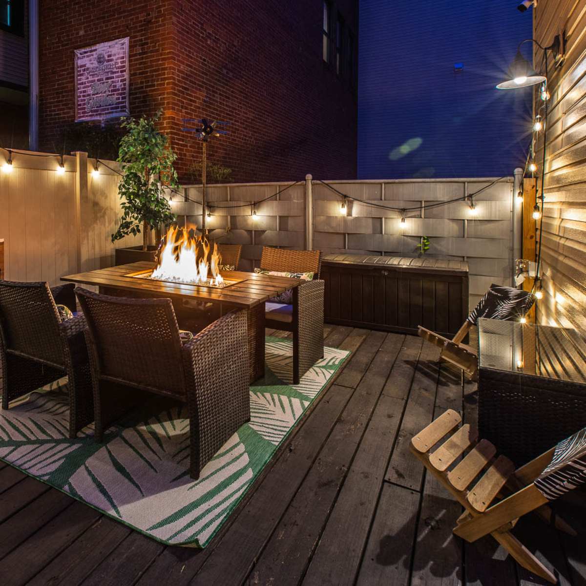 A cozy outdoor patio features a fire table surrounded by four wicker chairs, illuminated by string lights and set against a wooden deck.