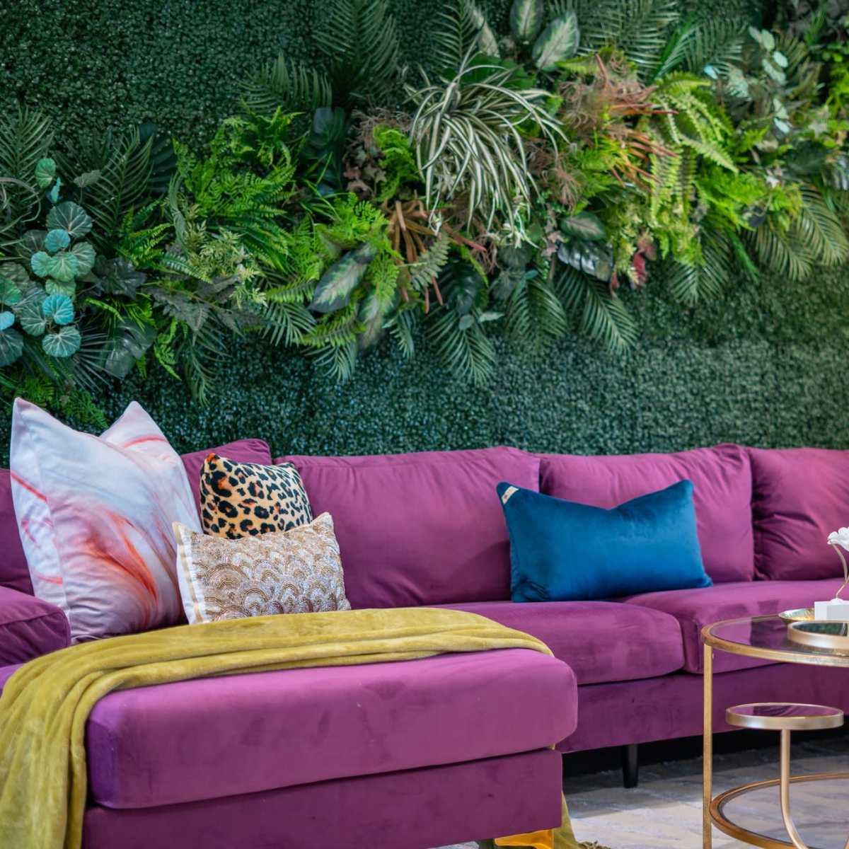 A modern living room features a purple sectional sofa, decorative pillows, a green vertical garden backdrop, and a round gold coffee table.