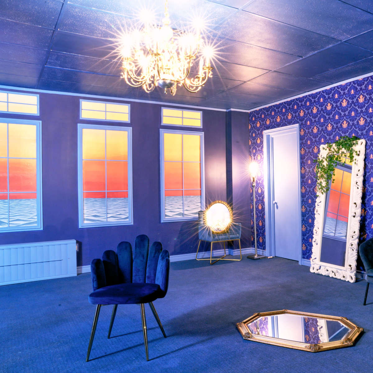 A room with purple wallpaper, a chandelier, a green velvet sofa, a decorative mirror, and large windows displaying a sunset view.