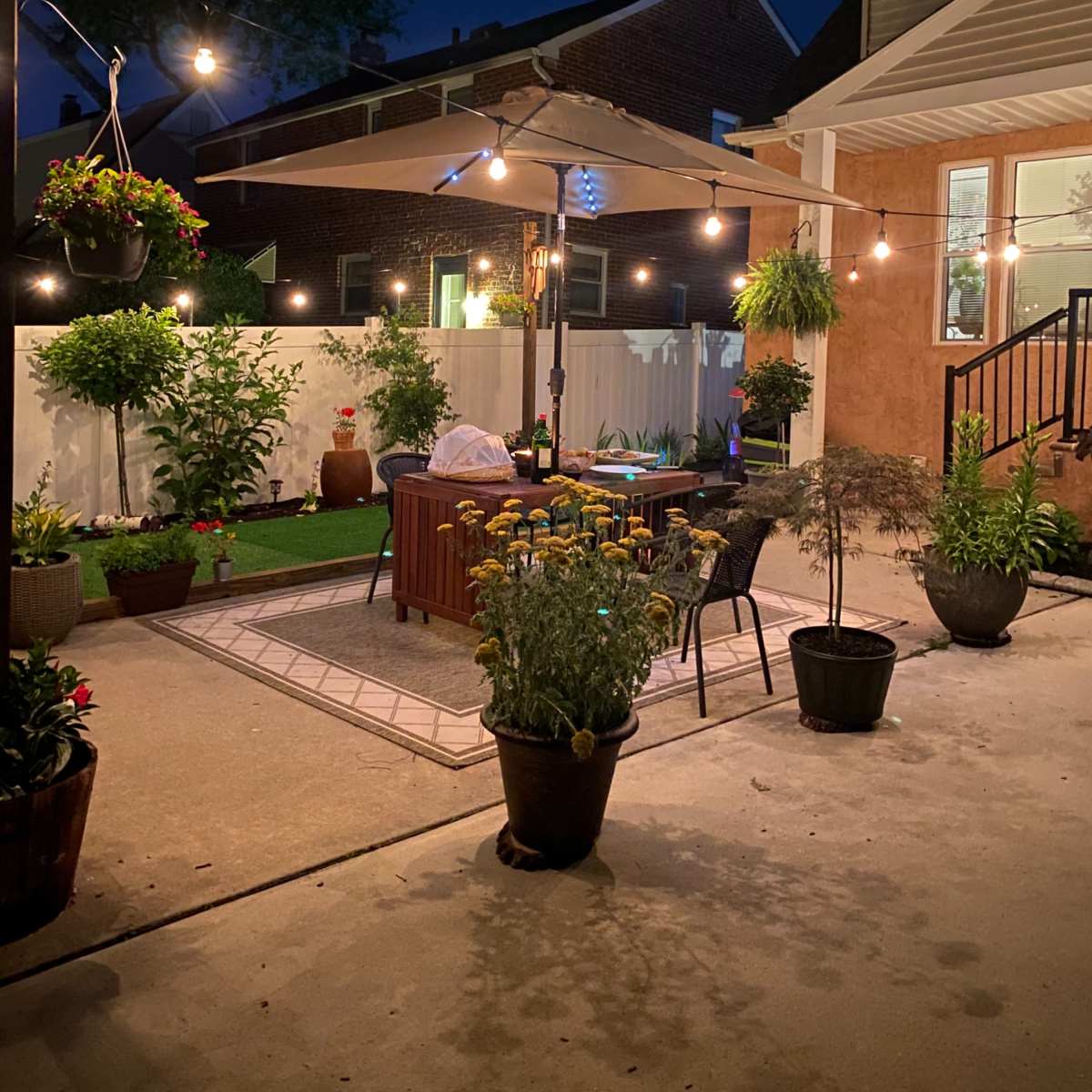 A backyard patio features a seating area under a large umbrella, surrounded by potted plants and string lights in the evening.