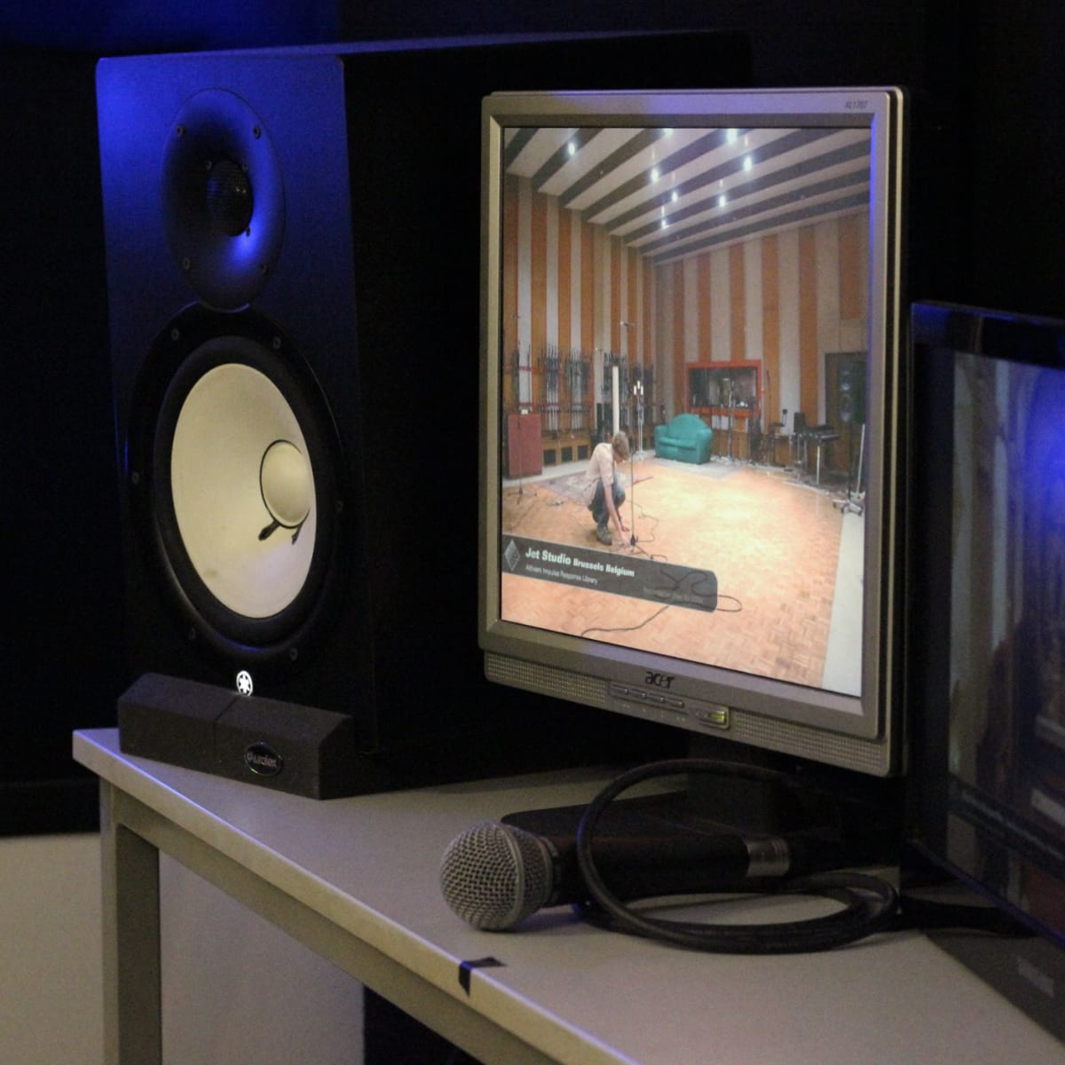 A computer monitor displays a video of a person in a recording studio, with audio speakers and a microphone nearby.