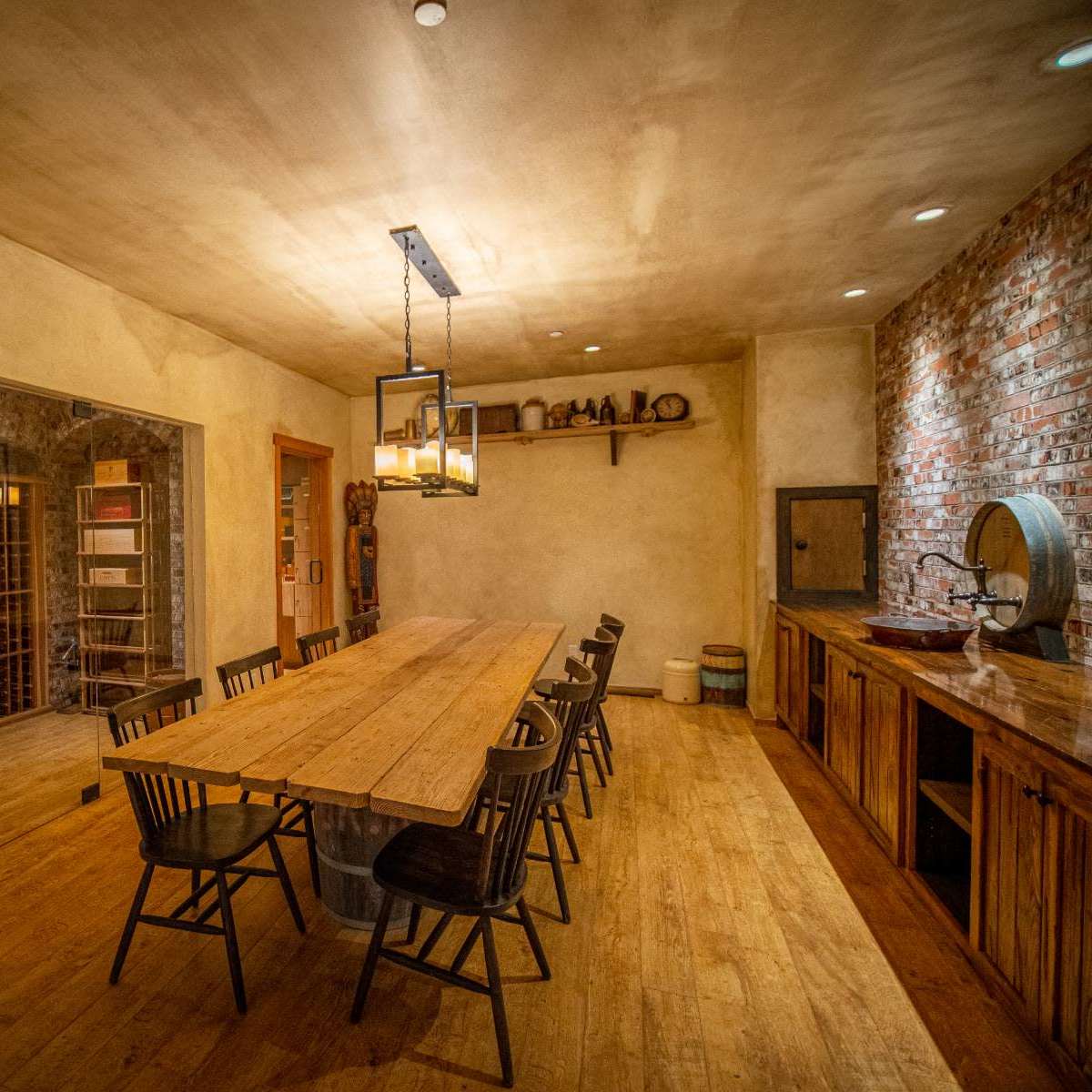A dining room with a long wooden table surrounded by chairs, a wine rack on one side, and a rustic sink area on the other.