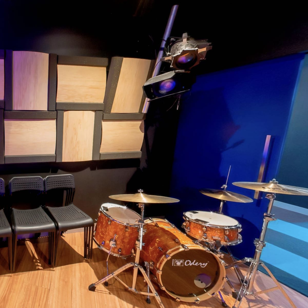 A music practice room with a drum set, a set of black chairs, and wooden sound panels on the wall.