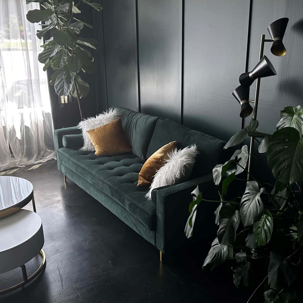 A green velvet sofa adorned with decorative pillows, a circular coffee table, and potted plants in a well-lit room with sheer curtains.