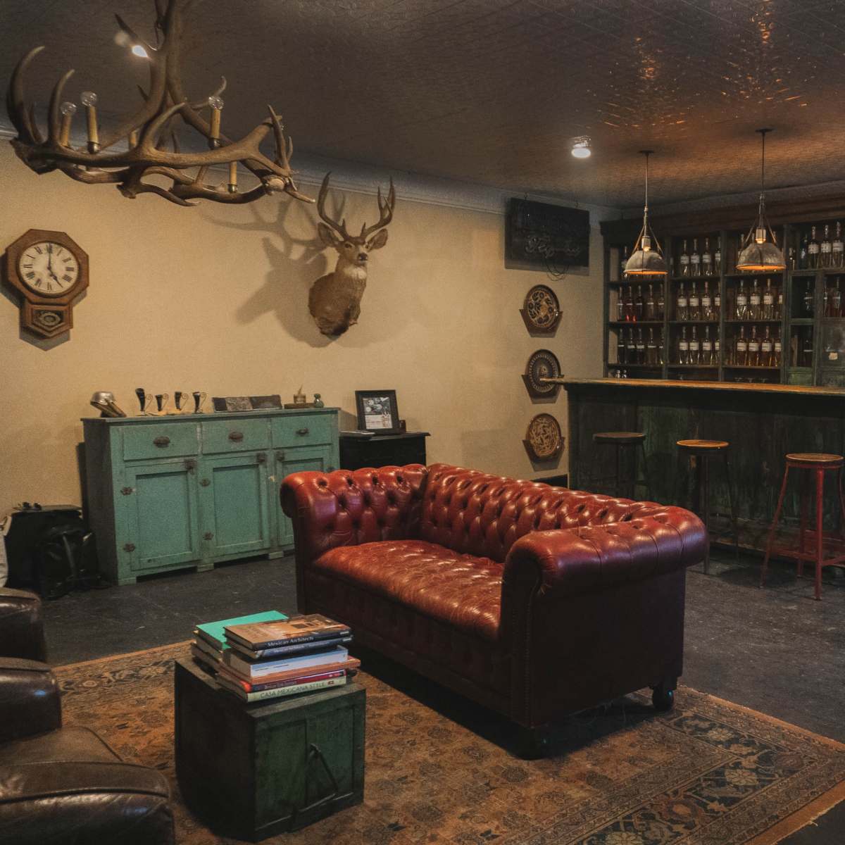 A cozy bar-style room featuring a red leather chesterfield sofa, a wooden bar stocked with bottles, and rustic decor including antlers and a clock on the wall.