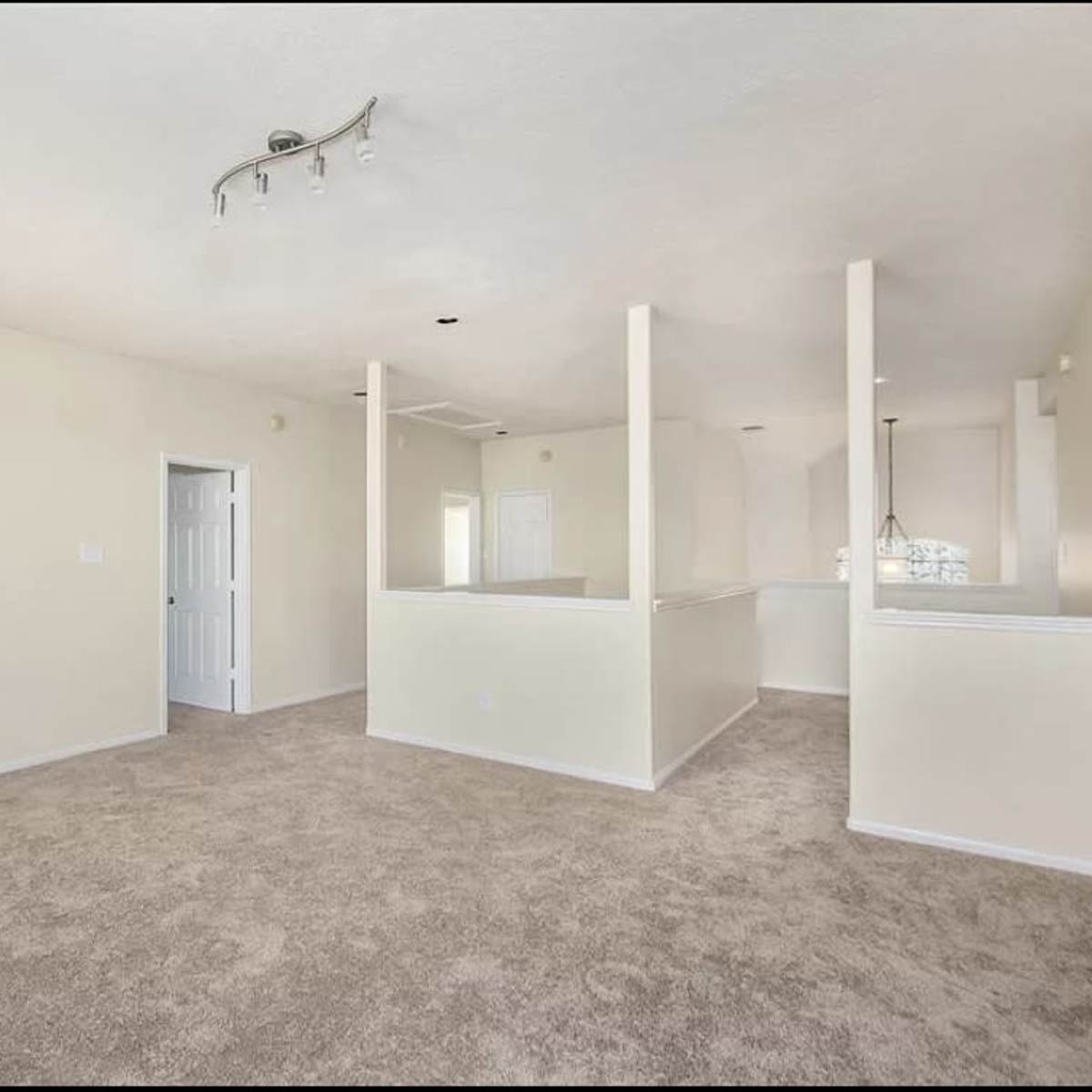A spacious, carpeted living area with light-colored walls and a ceiling fixture, flowing into an open layout that includes a kitchen area.
