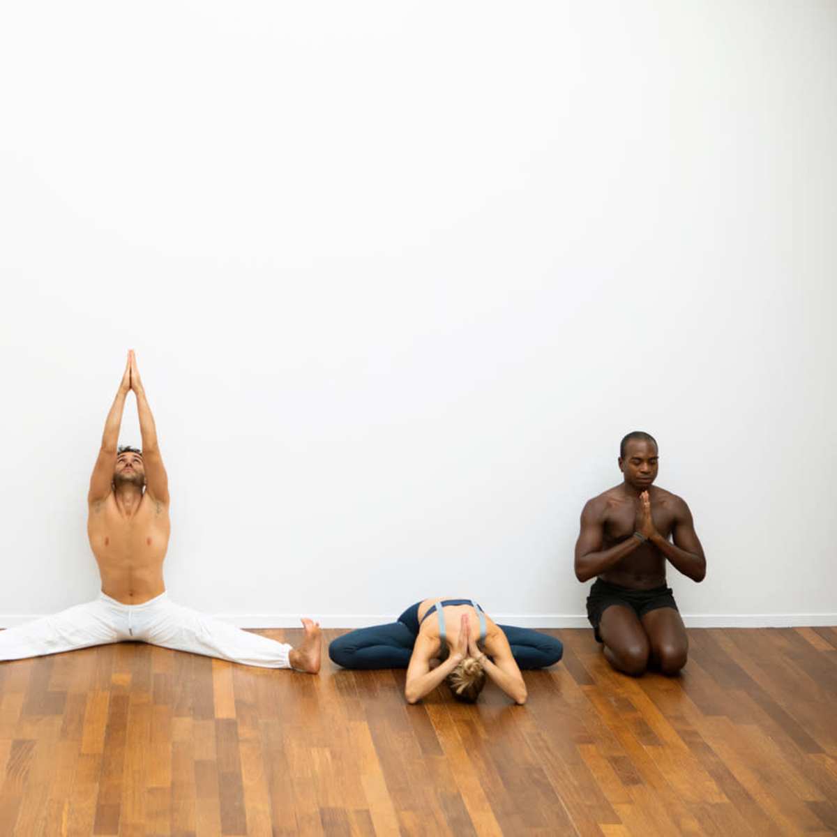 Four individuals practice yoga on a hardwood floor, each in a different pose against a plain white wall.