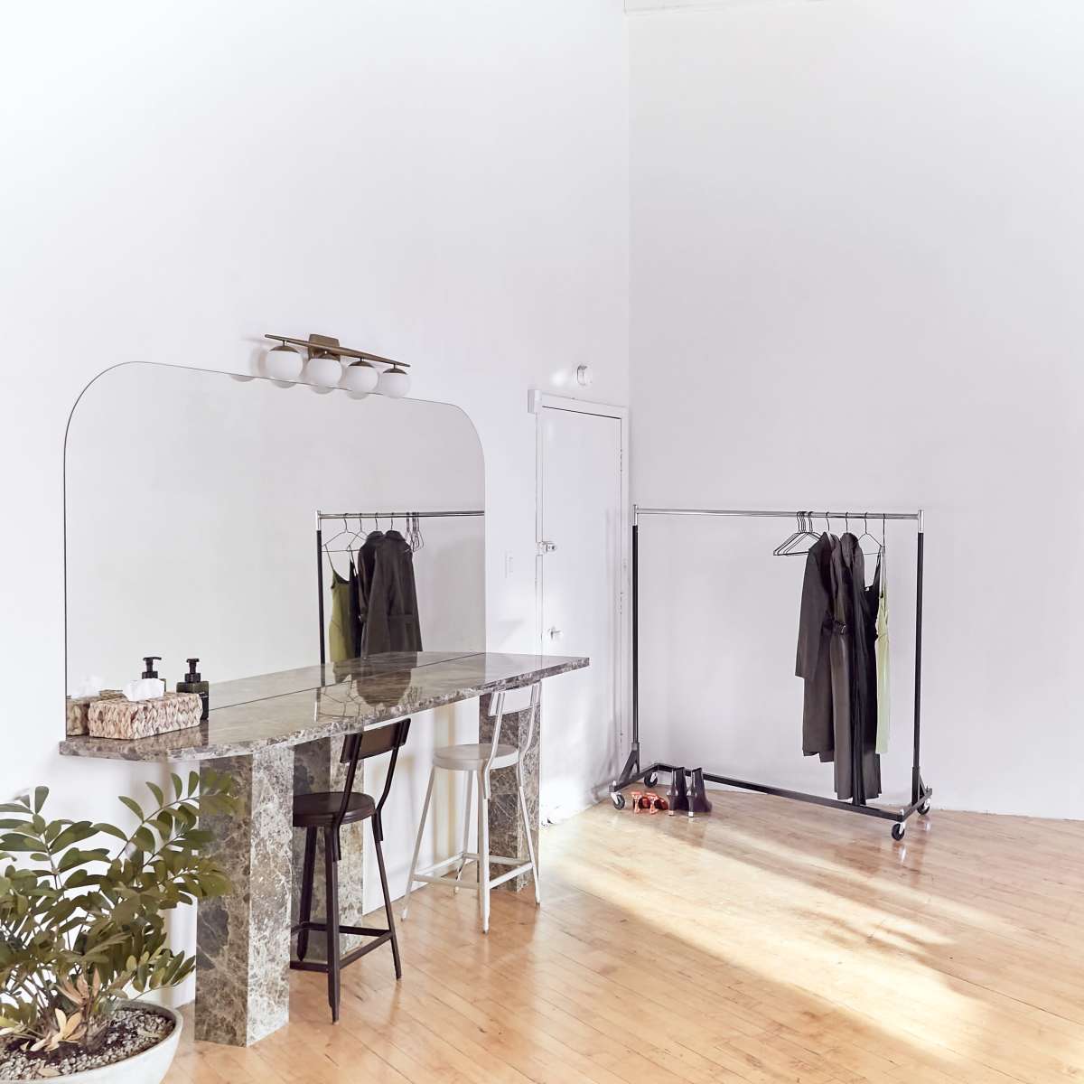 A minimalist dressing area features a mirror, a marble table, a clothing rack with garments, and a small plant in a light-filled room with wooden flooring.
