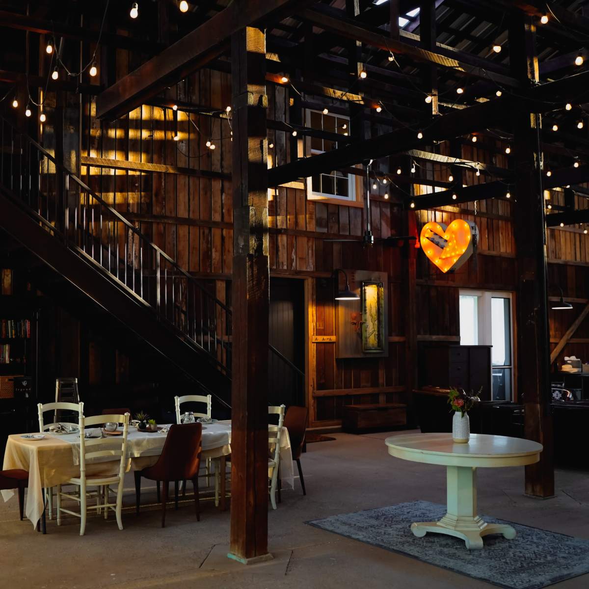The interior of a rustic, wooden barn features a dining area with a table and chairs beneath string lights and a heart-shaped decor piece.