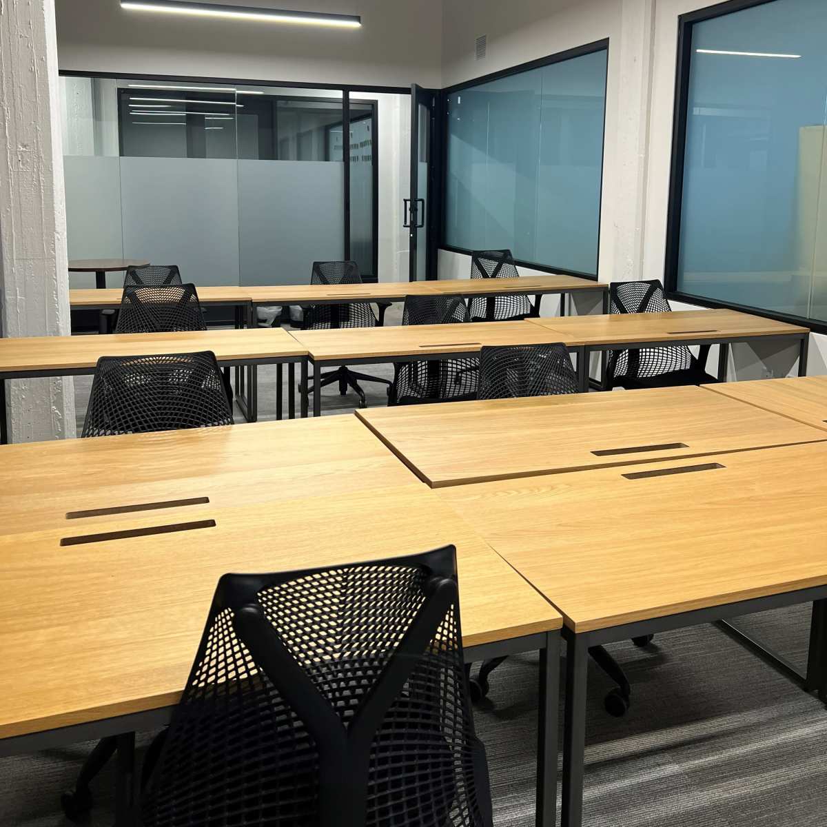 An empty modern office space with multiple wooden desks and mesh chairs arranged in a grid pattern, separated by glass partitions.