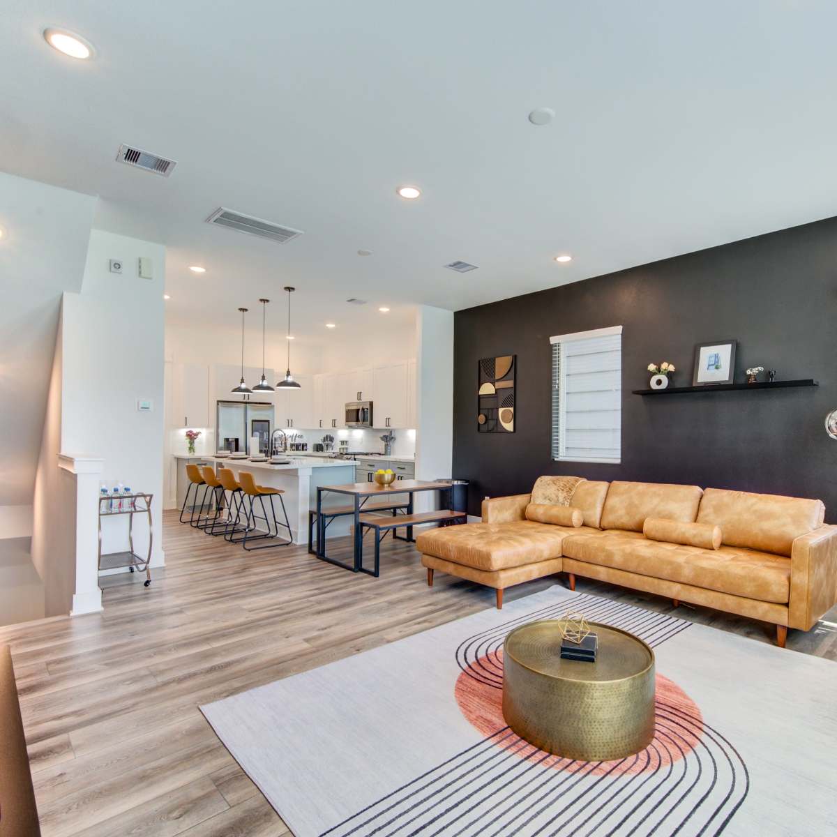 A modern living room with a brown leather sectional sofa, a yellow accent chair, a circular coffee table, and a kitchen area in the background.