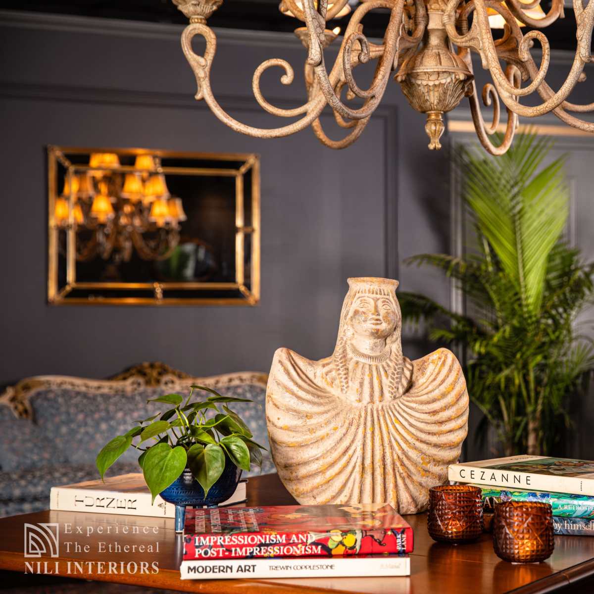 A well-decorated room features a chandelier above a wooden table displaying a decorative vase, books, and a small plant, with upholstered furniture and artwork in the background.