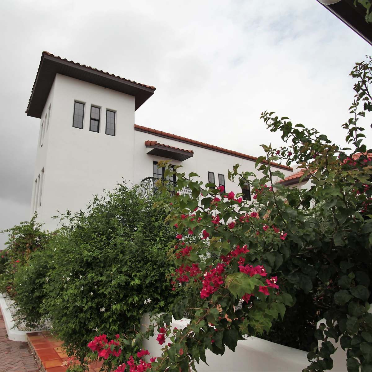 A white stucco building with a tile roof is situated alongside a pathway adorned with vibrant pink bougainvillea.