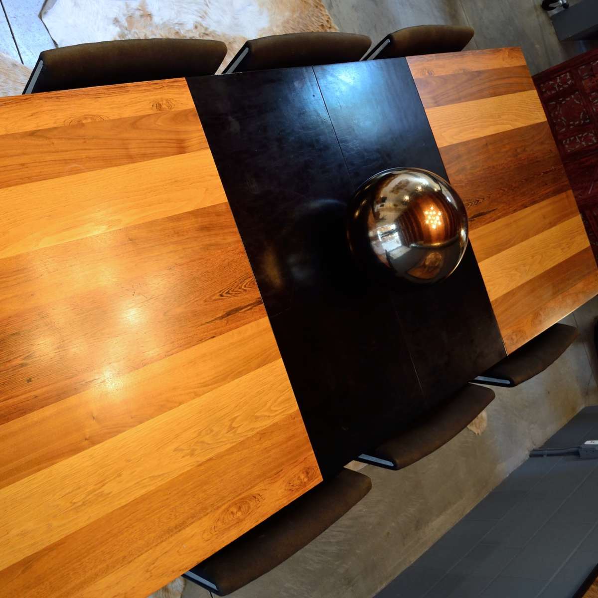 A long dining table made of alternating light and dark wooden planks, surrounded by several chairs, with a round metallic centerpiece.