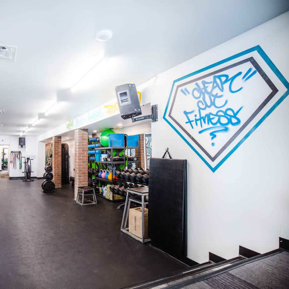 A gym interior featuring weightlifting equipment, a wall adorned with graffiti, and neatly organized fitness gear on shelves.