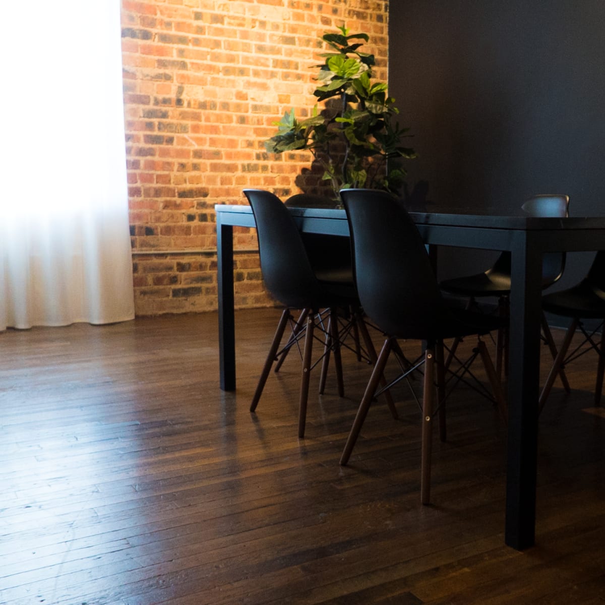 A wooden dining table with black chairs sits in a room featuring exposed brick walls and a large window partially covered by white curtains.